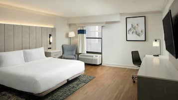 A hotel room with a large bed, a desk, a chair, a TV, a window with curtains, and a wall-mounted artwork.