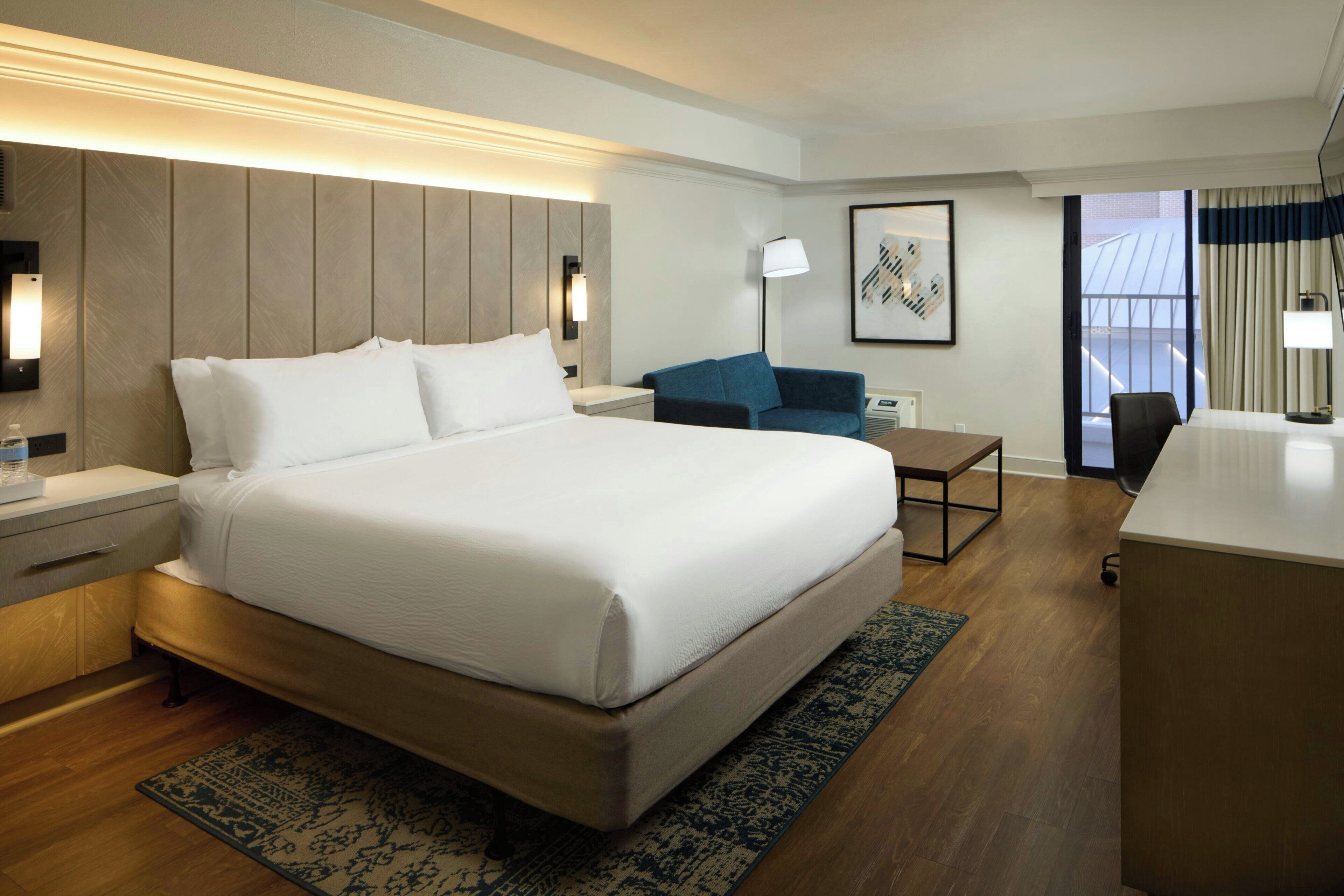 A hotel room with a large bed, a desk, a chair, a small table, and a sofa.