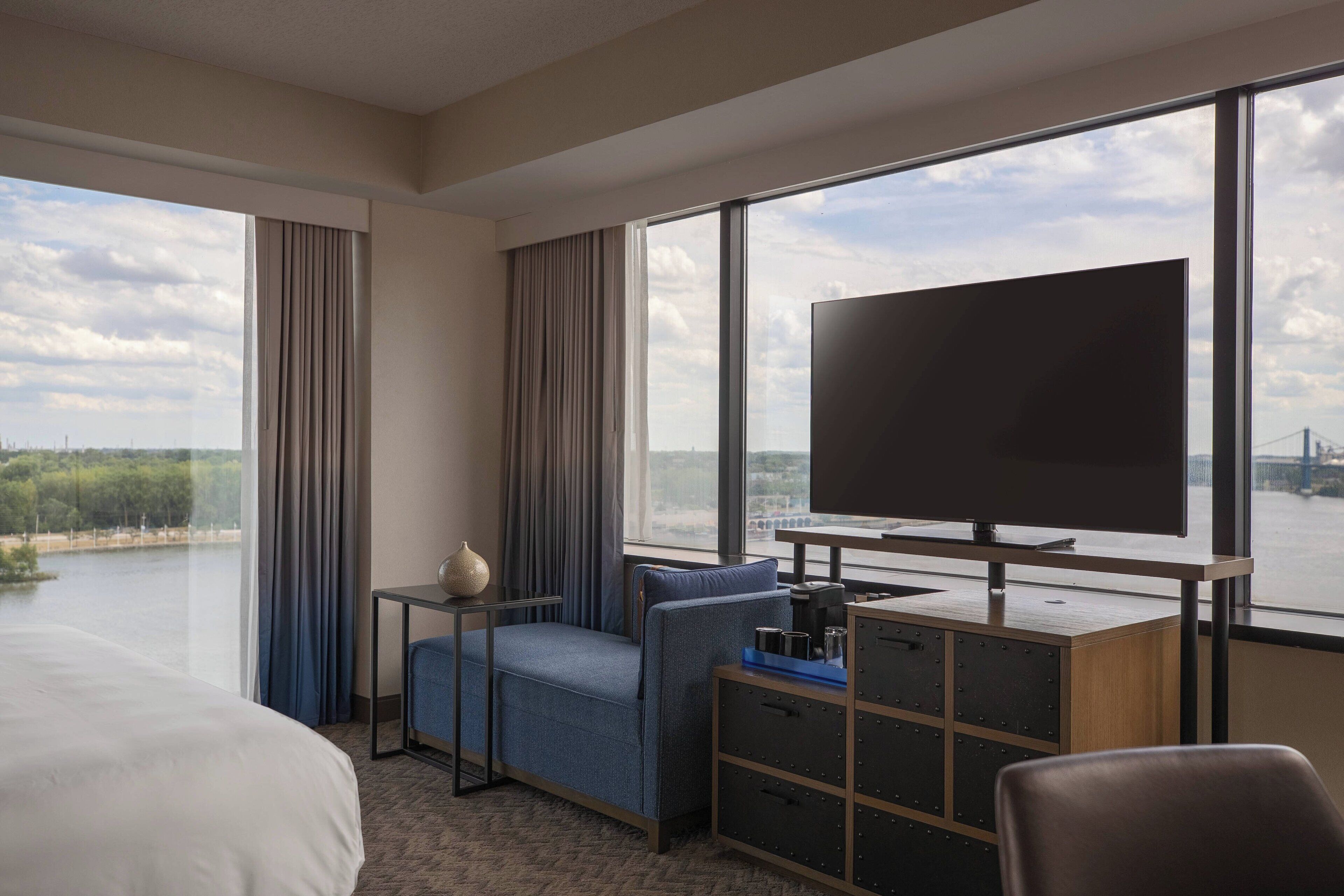 A hotel room with a large bed, a TV on a wooden cabinet, a desk with a chair, and a view of a body of water and a bridge.