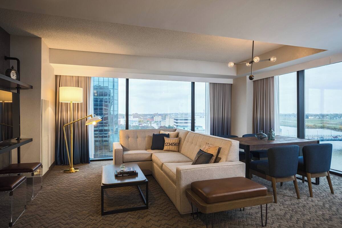 A modern hotel room with a large sofa, a dining table, and chairs. There is a lamp, a clock, and a view of the city through large windows.