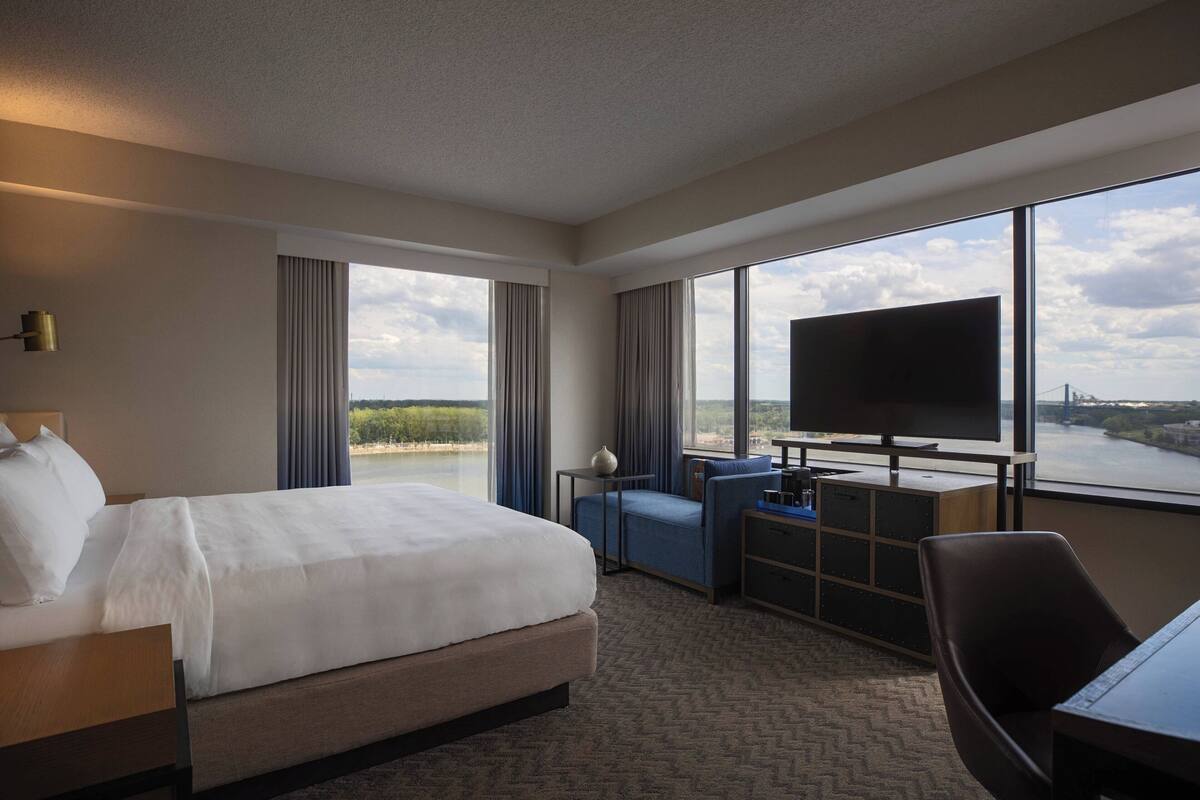 A hotel room with a large bed, a desk with a flat-screen TV, a chair, and a view of a river and bridge.