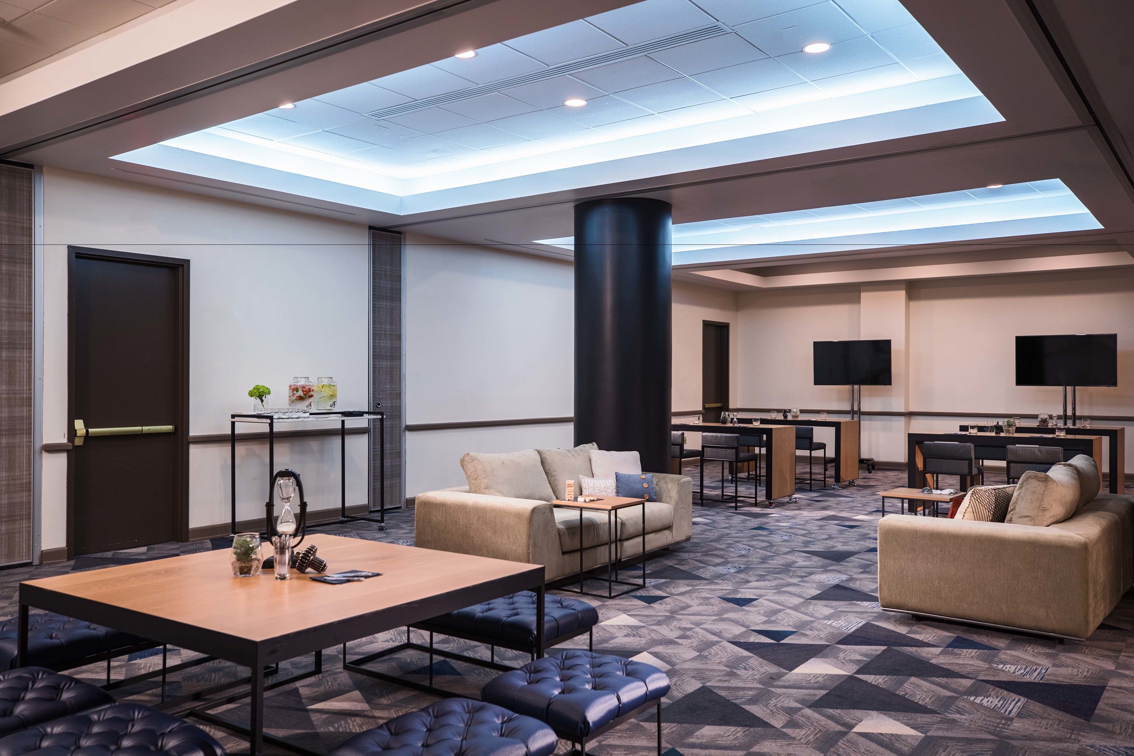 Un hall d’hôtel moderne avec un plafond haut, un espace salon central agrémenté de canapés et d’une table, une grande colonne et un téléviseur à écran plat fixé au mur.