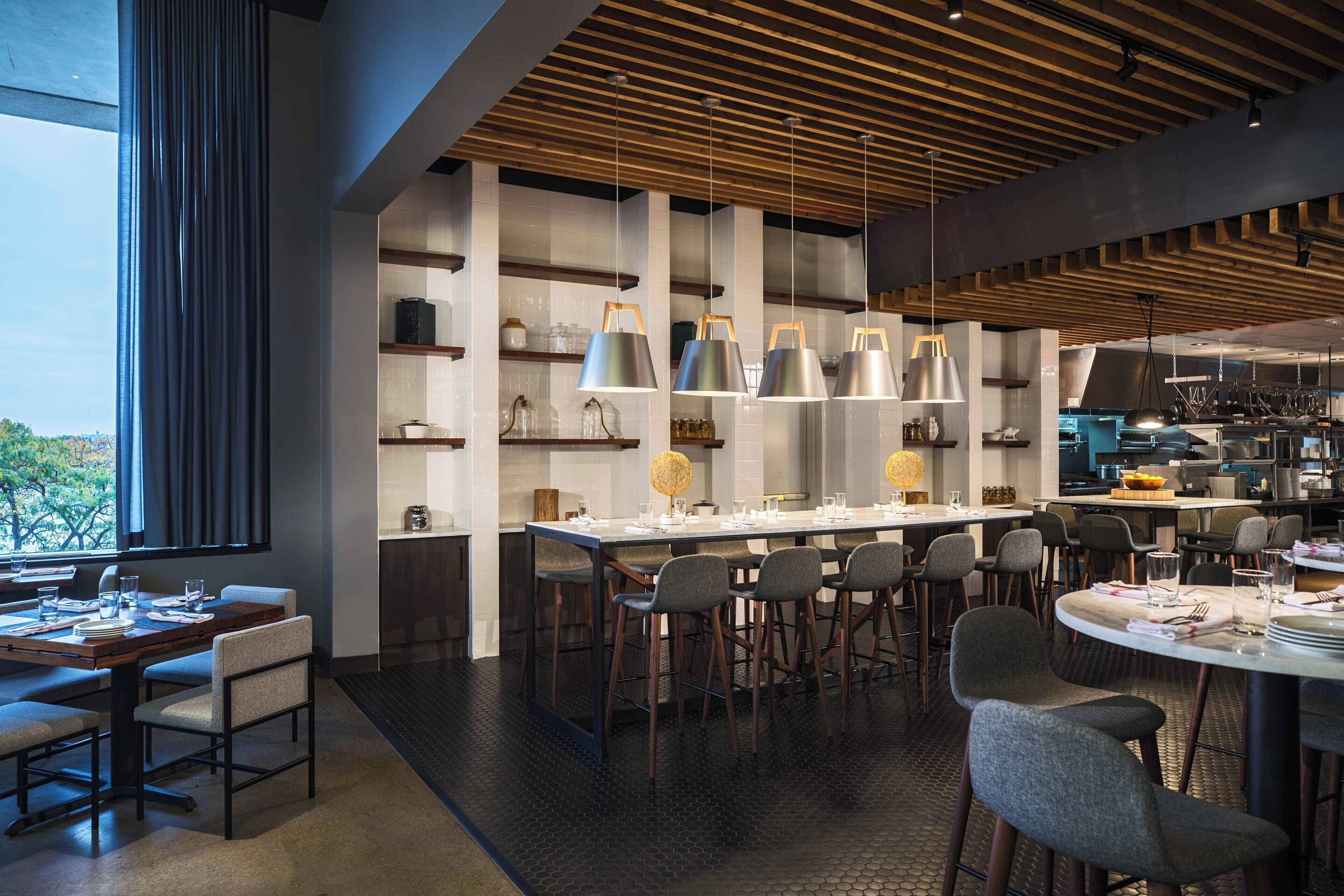 A modern restaurant with a long communal table, individual seating, and a kitchen area visible in the background.