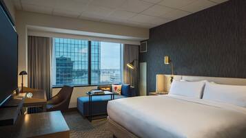 A hotel room with a large bed, a desk with a computer, a chair, a small table, a lamp, and a view of the city through large windows.