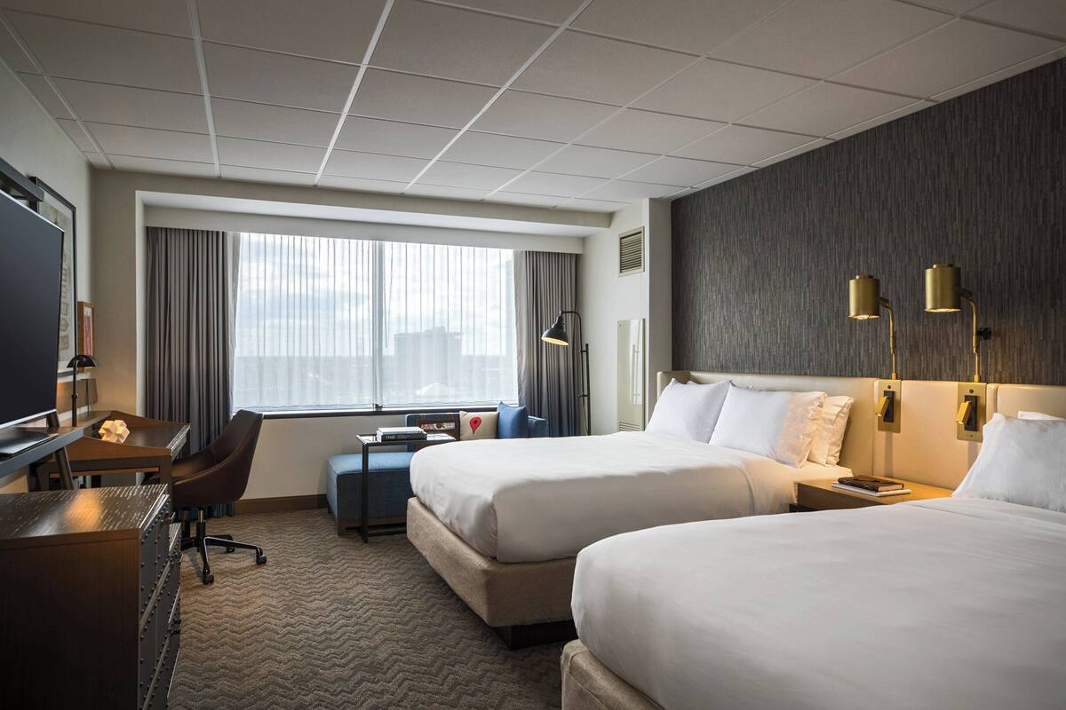 A hotel room with two beds, a desk, a chair, a TV, and a large window with curtains.