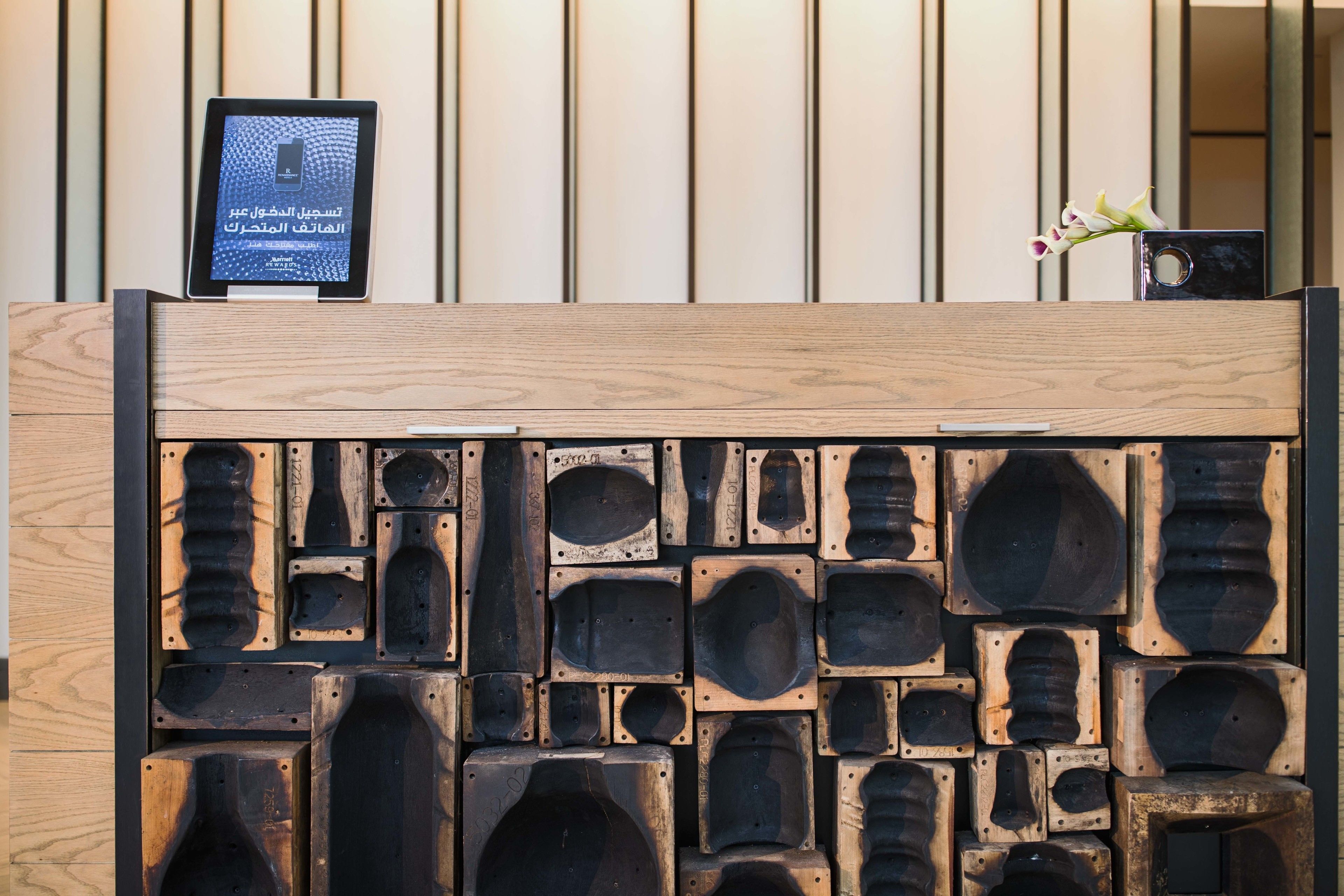 A wooden reception desk with a tablet displaying Arabic text, a vase with flowers, and a variety of wooden planters.