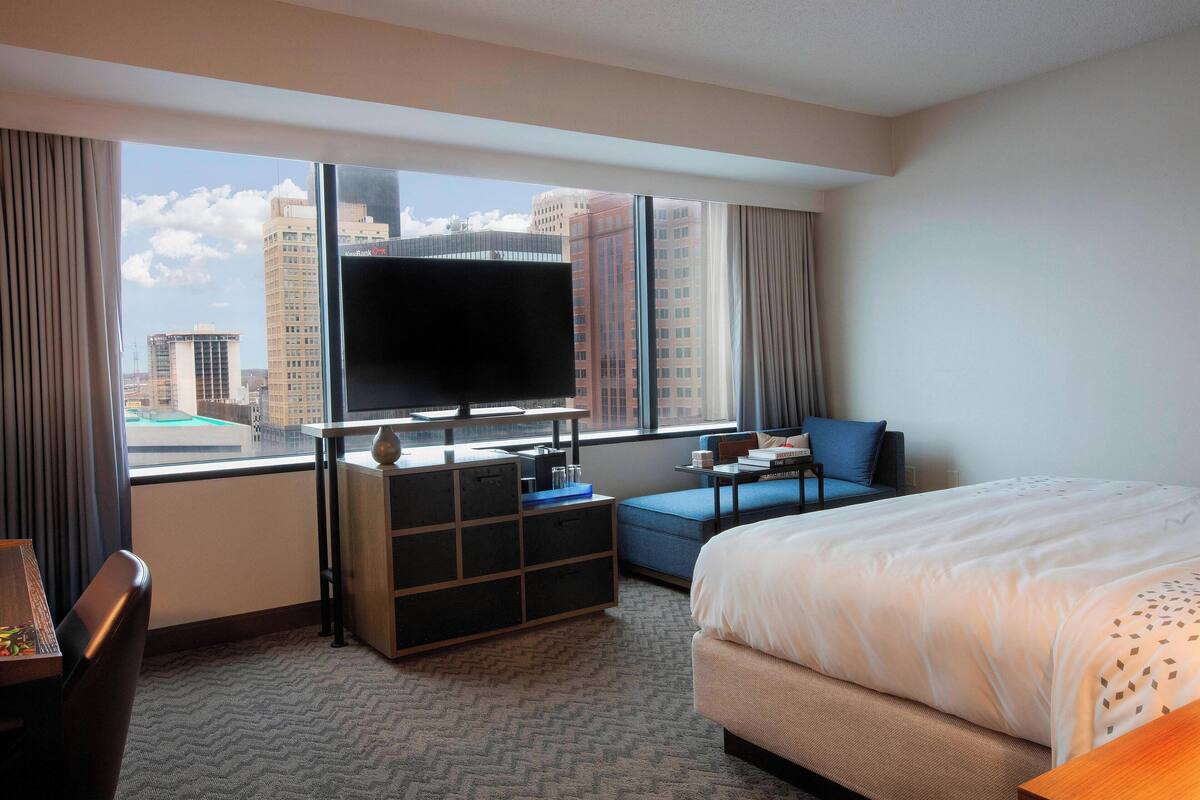 A hotel room with a large window, a bed, a TV, a desk, and a sofa.