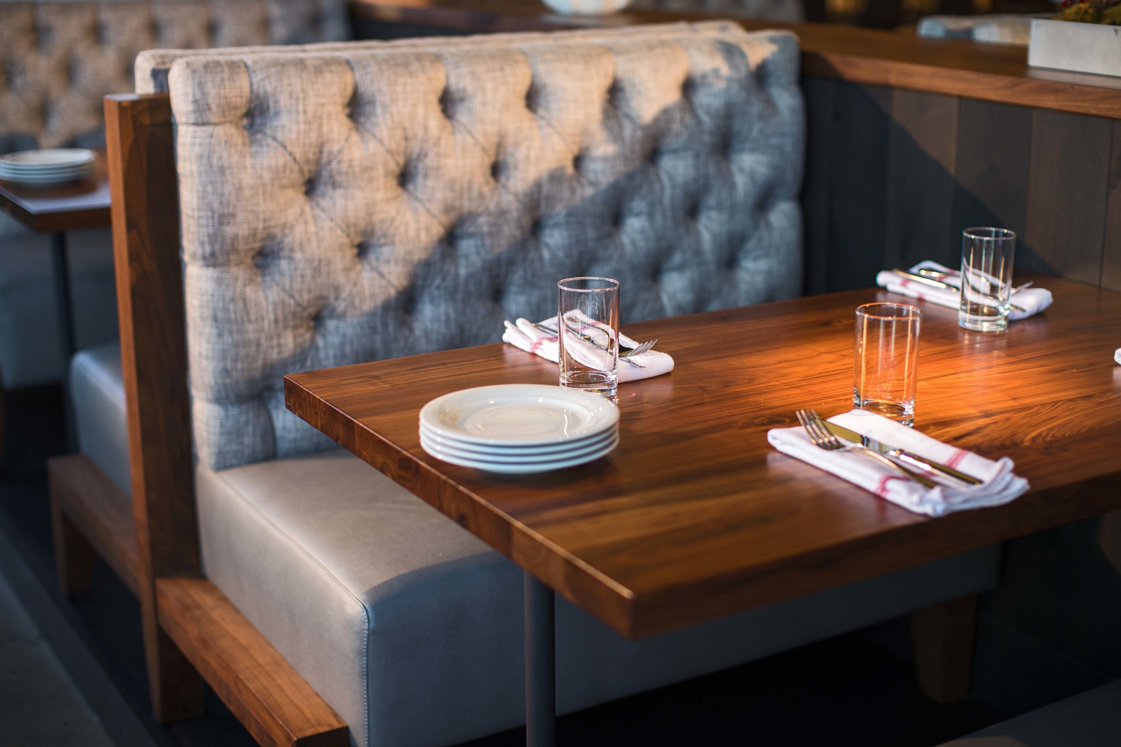 A dining area with a wooden table set for two, a tufted upholstered bench, and a stack of plates on the table.