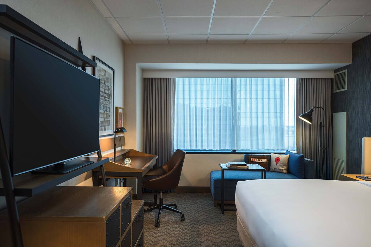 A hotel room with a bed, a desk with a chair, a television, and a window with curtains.