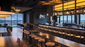 Eine Bar mit einer langen Theke, Barhocker und Blick auf die Stadt durch große Fenster.