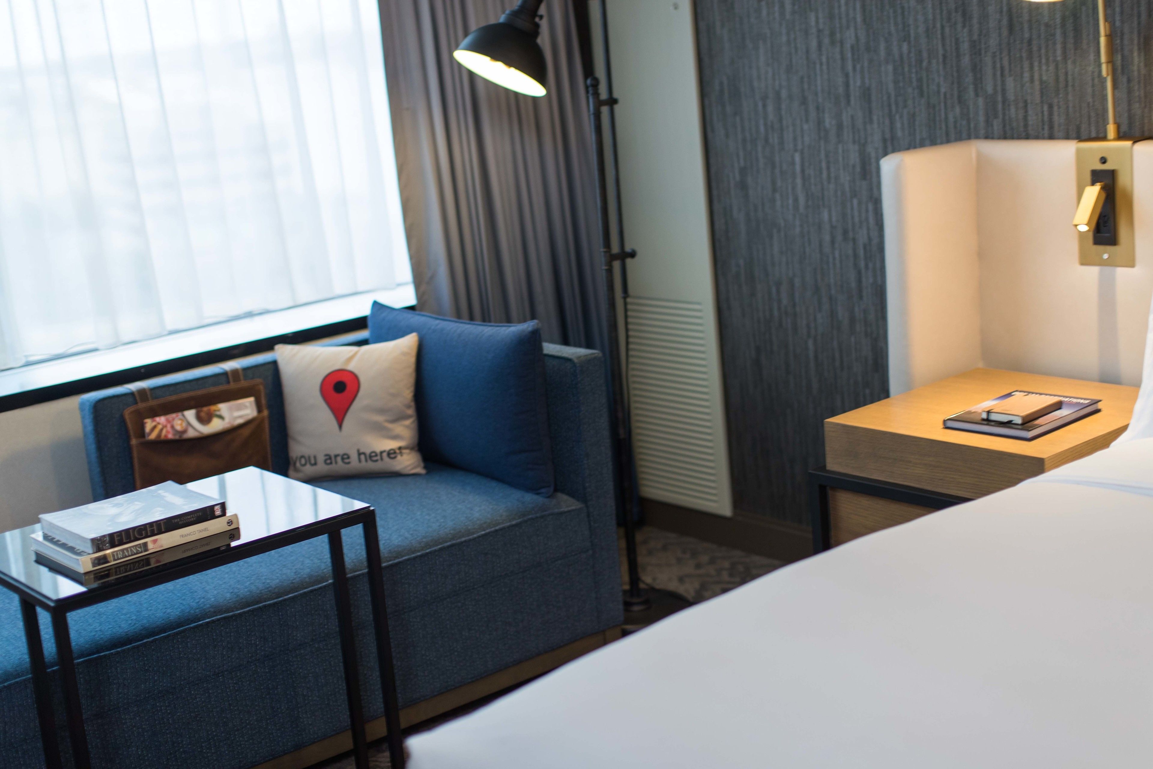 A hotel room with a sofa, a small table with books, a lamp, and a bedside table with a book.