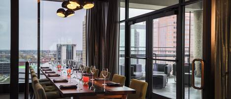 Un comedor con una mesa larga, varias sillas y vistas a un paisaje urbano a través de amplios ventanales.