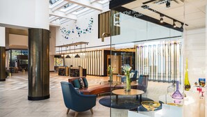 Eine großzügige Lobby mit modernem Design, ausgestattet mit einer Sitzecke mit Sofas und einem Couchtisch, einer Glasfront mit einem Schild sowie einer hohen Decke mit Oberlichtern.