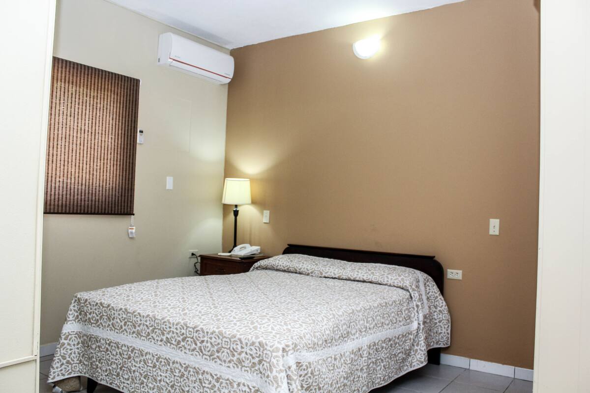 A hotel room with a bed, bedside lamp, and an air conditioning unit.