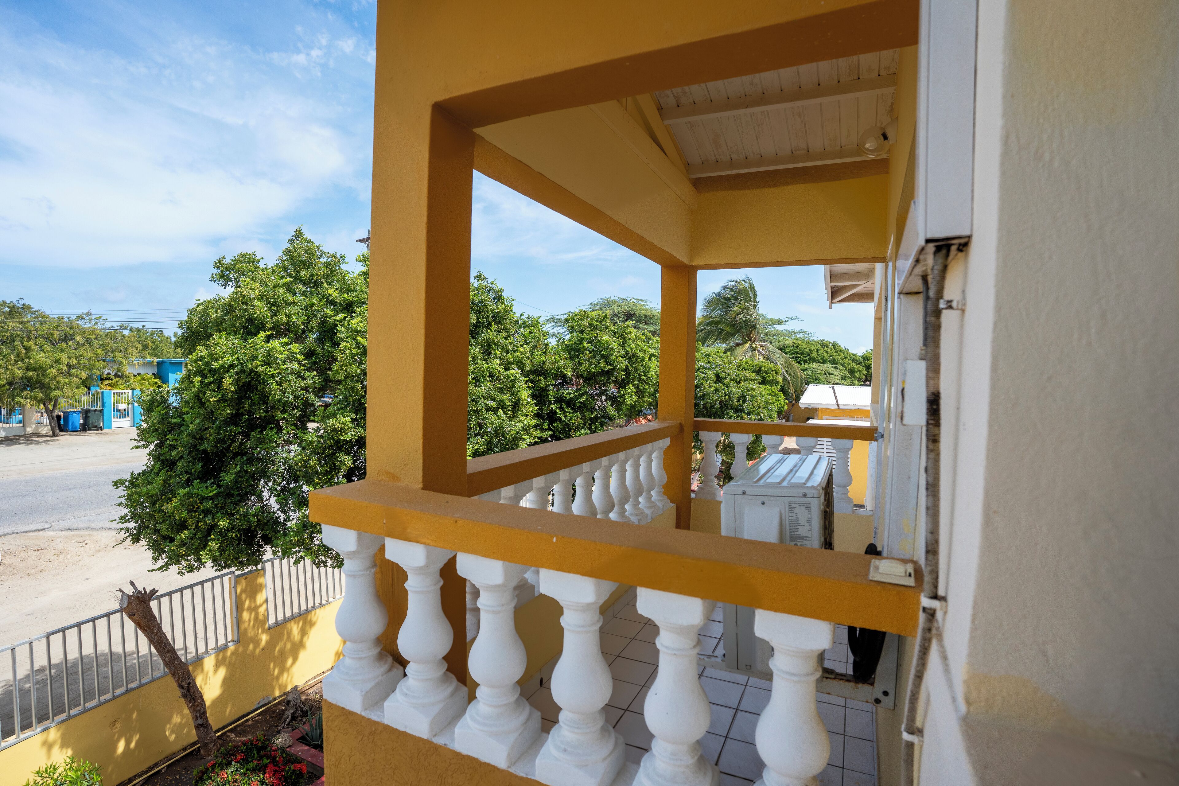 A balcony with white balustrades and a yellow wall, overlooking a street with trees and buildings.