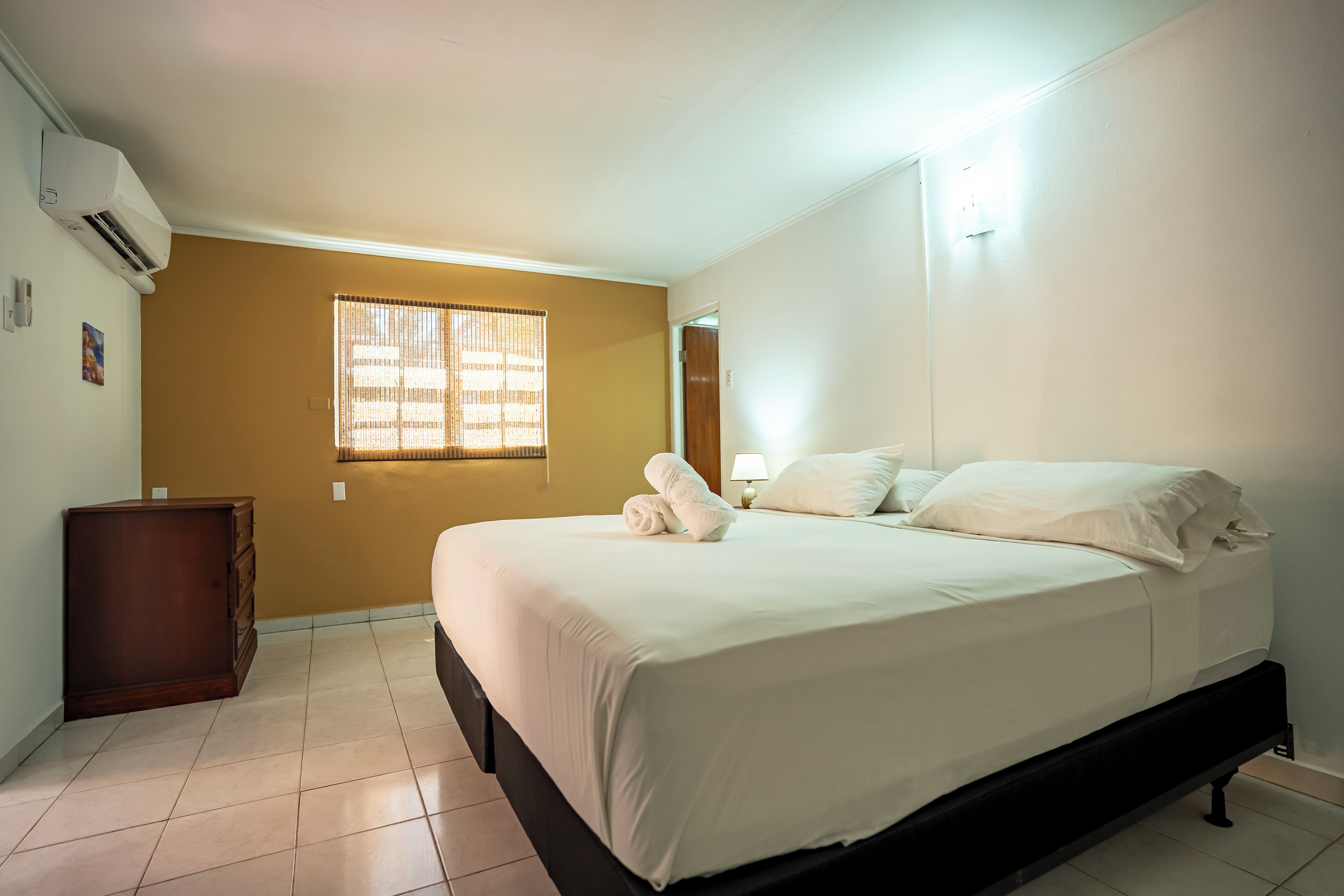 Un dormitorio con una cama, un aparador de madera, una ventana con persianas y un equipo de aire acondicionado.