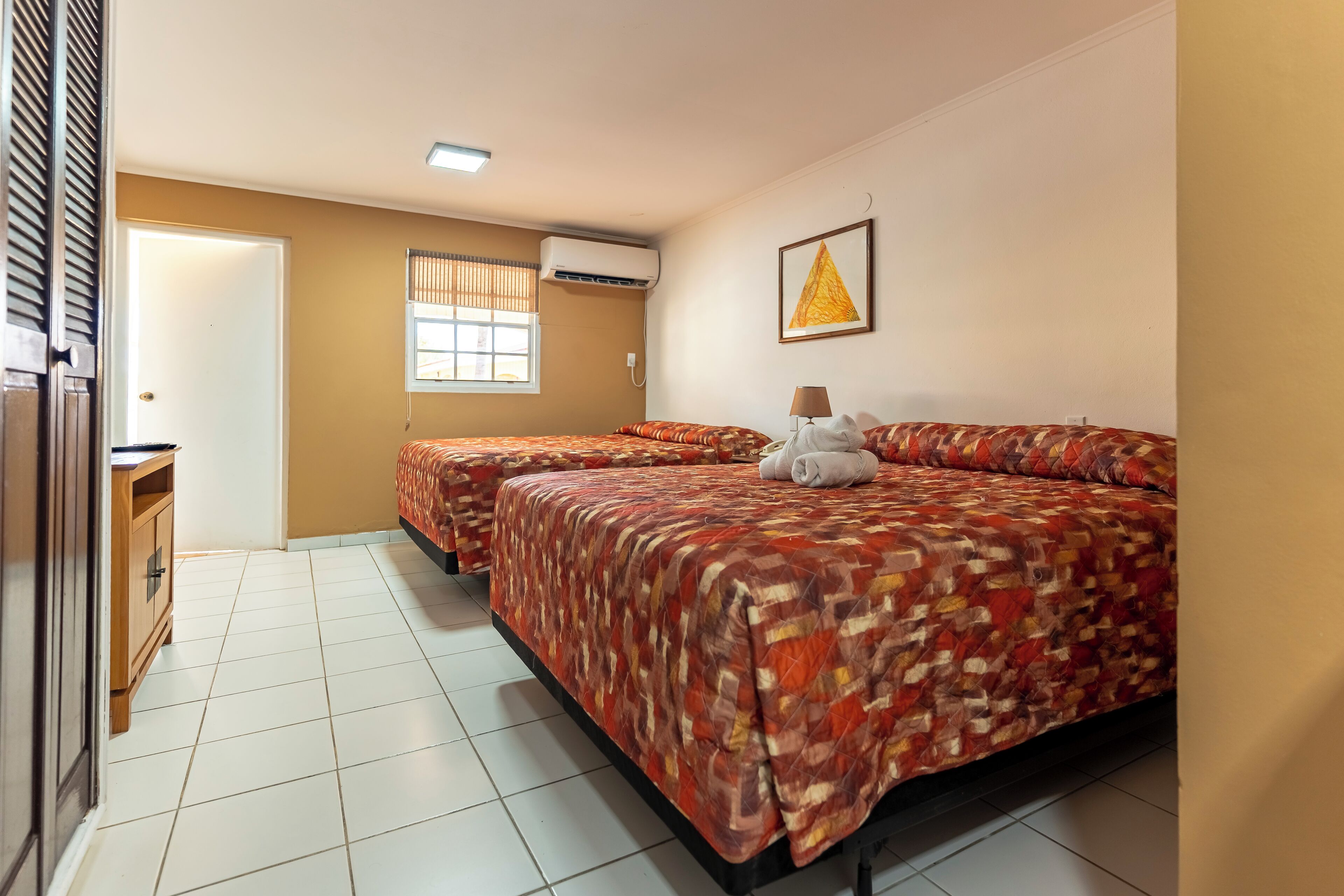 Habitación de hotel con dos camas, una ventana con persianas, un equipo de aire acondicionado, un cuadro enmarcado en la pared y una puerta.