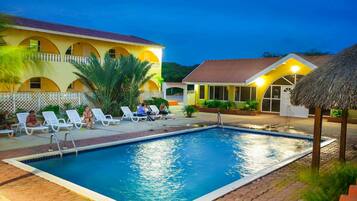 A swimming pool with lounge chairs, a thatched-roof structure, and a building with arched windows.