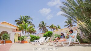 A pool area with lounge chairs, palm trees, and a building with a yellow facade. - Coconut Inn (Noord)