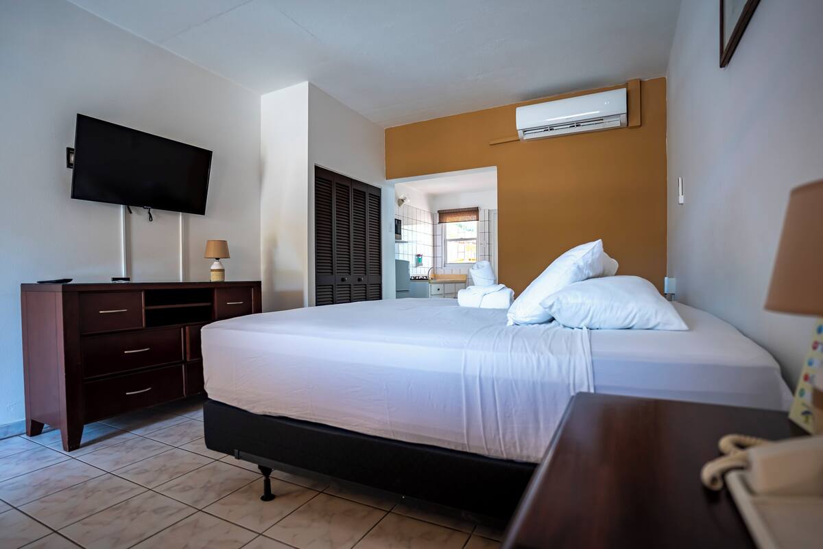 A hotel room with a large bed, a TV mounted on the wall, a desk with a lamp, and an air conditioning unit.