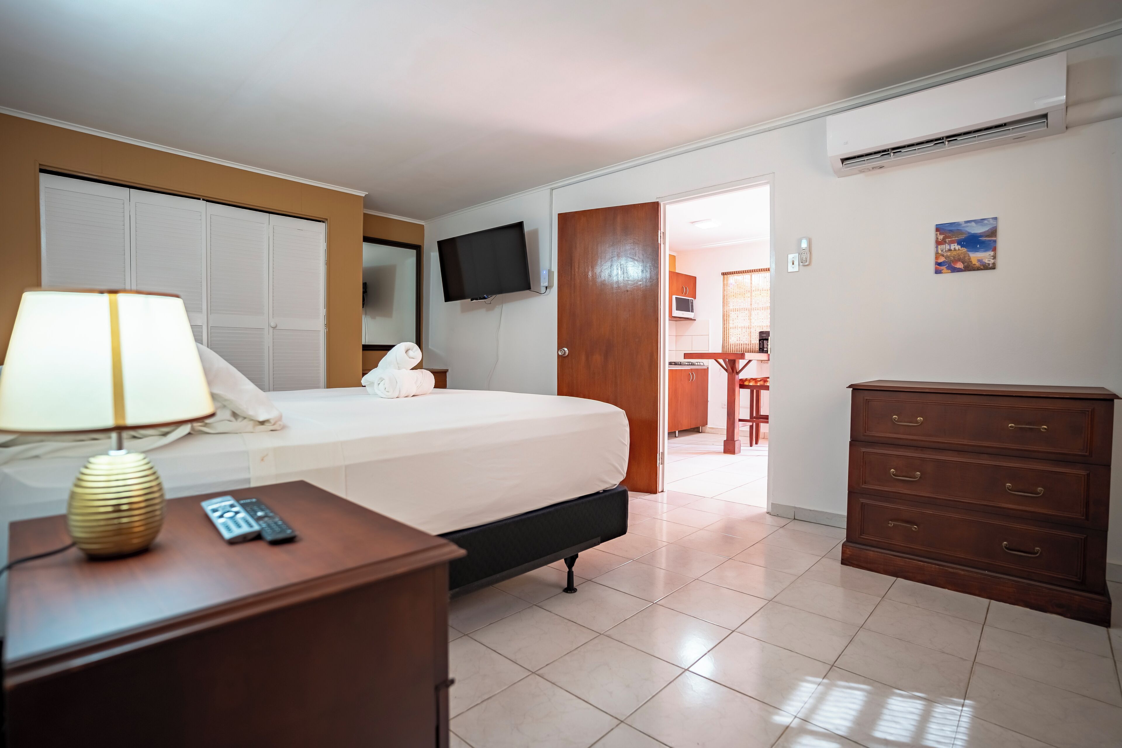 Une chambre d’hôtel comprenant un lit, une table de chevet avec une lampe, une commode, un téléviseur fixé au mur et un coin cuisine visible à travers une porte ouverte.
