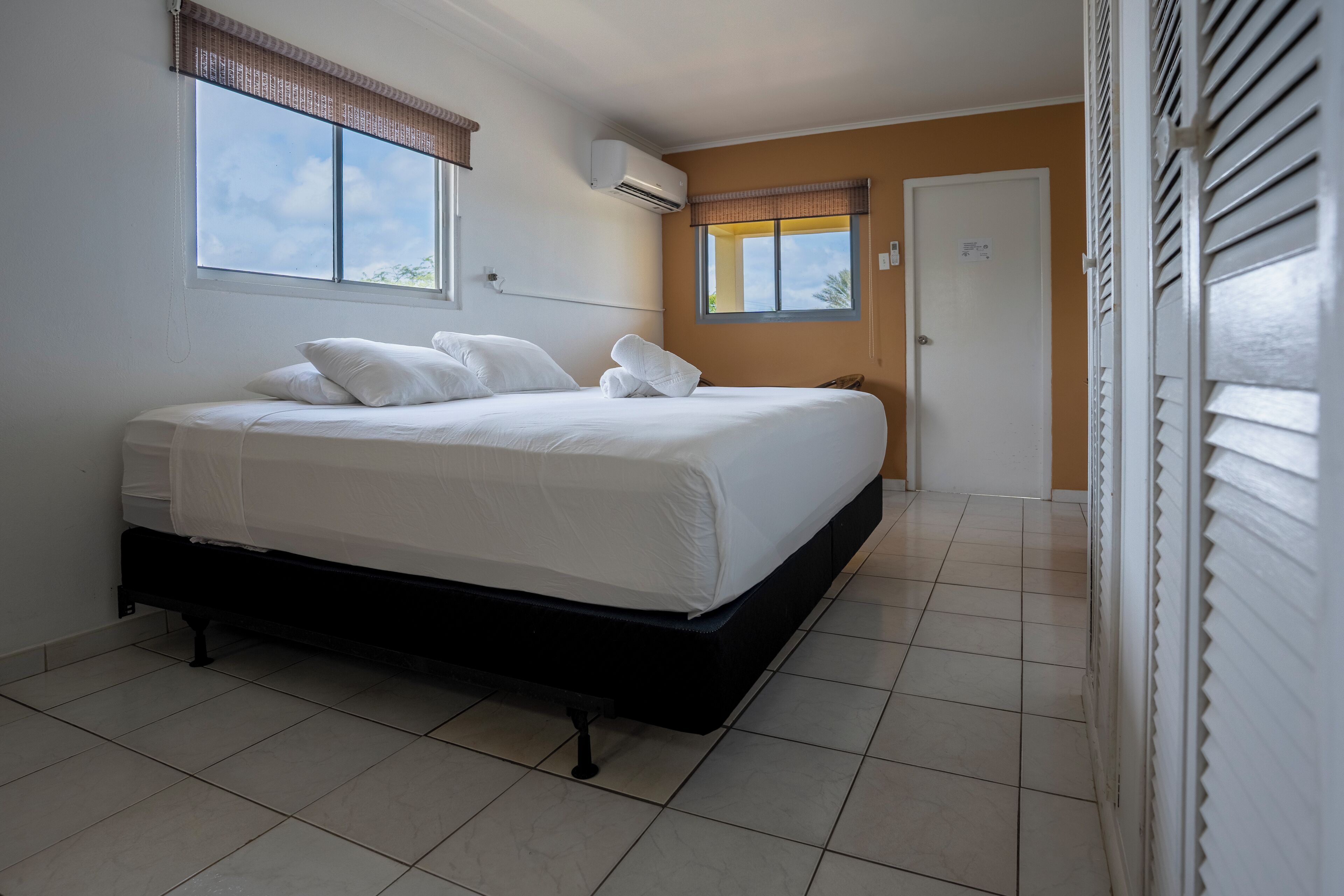 A bedroom with a bed, an air conditioner, a window with blinds, and a door.