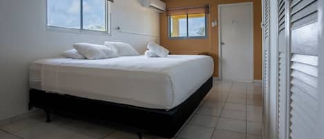A bedroom with a bed, an air conditioner, a window with blinds, and a door.