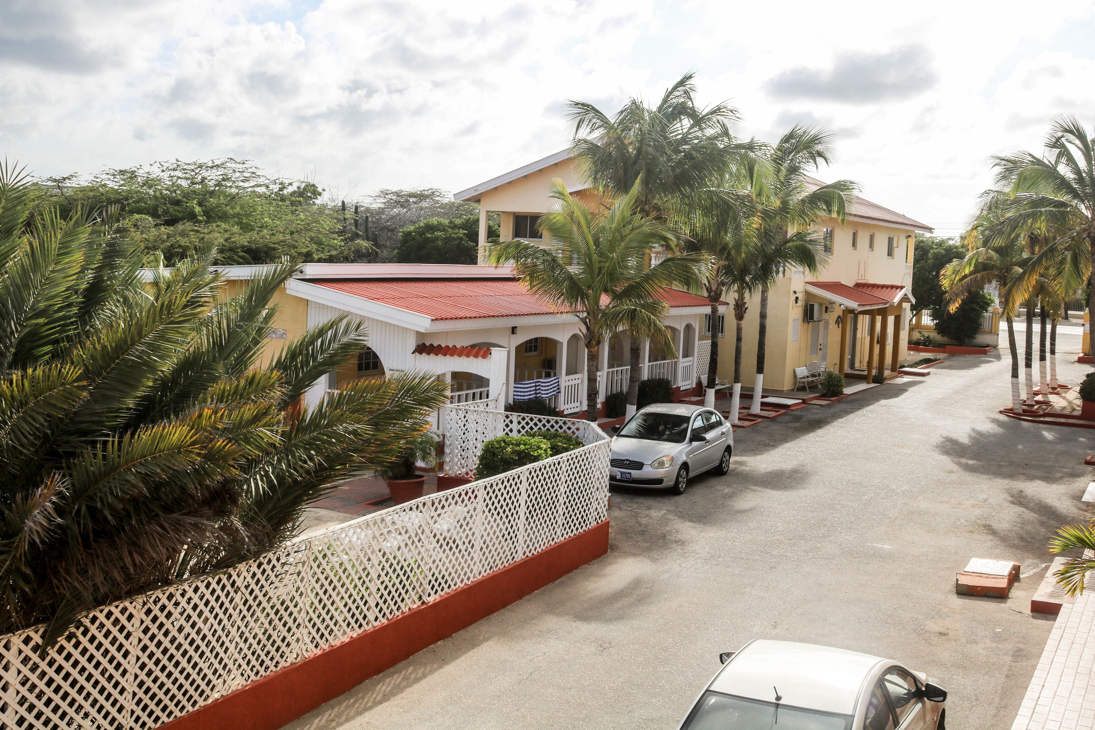 Vista de la calle con un edificio blanco de techo rojo, palmeras y coches estacionados.