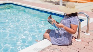 Une femme est assise au bord d’une piscine, un livre à la main, et porte un chapeau et une robe à motifs.