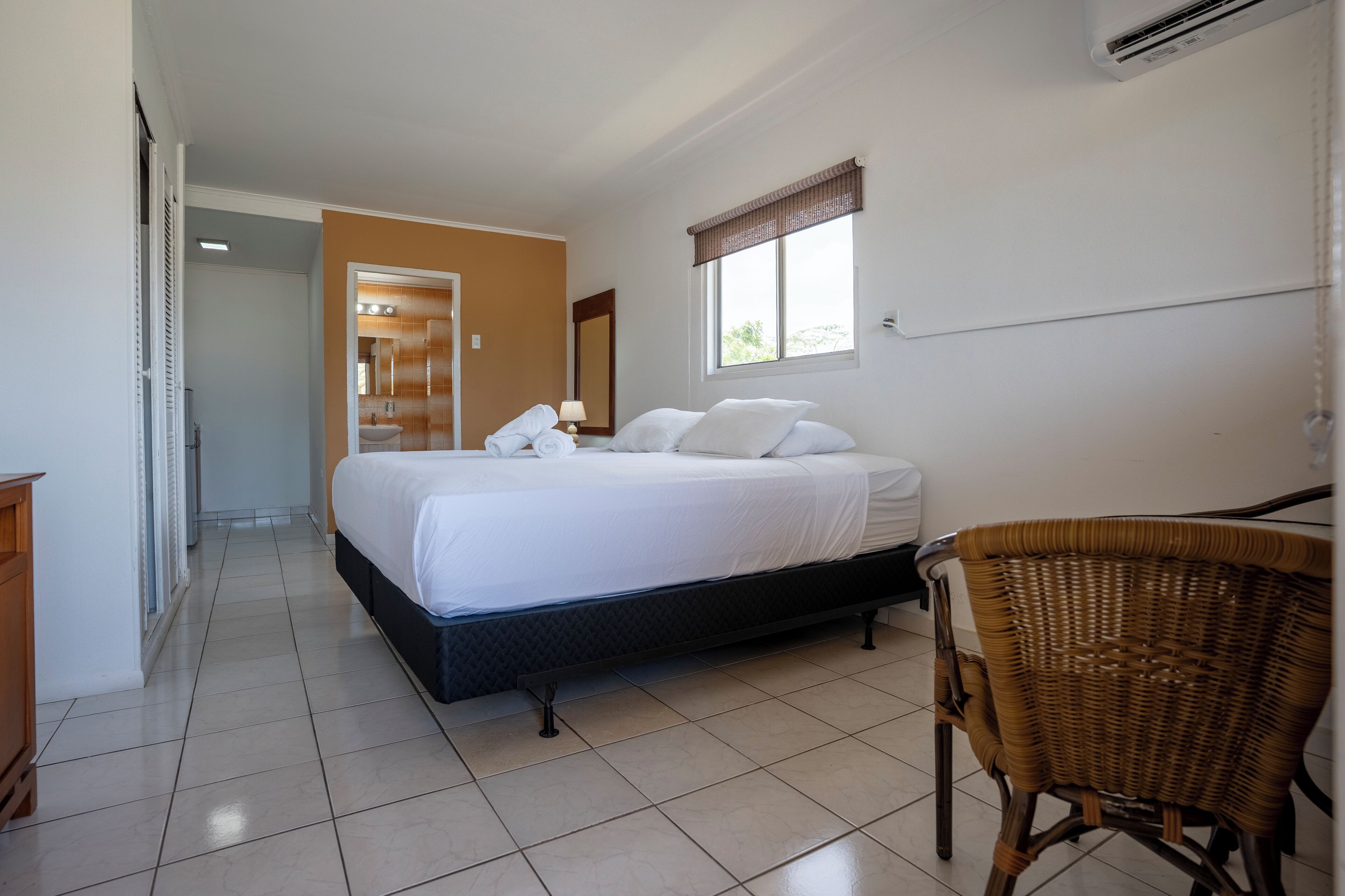 Un dormitorio con cama, una silla, un ventanal y un baño visible al fondo.