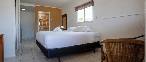 Un dormitorio con cama, una silla, un ventanal y un baño visible al fondo.