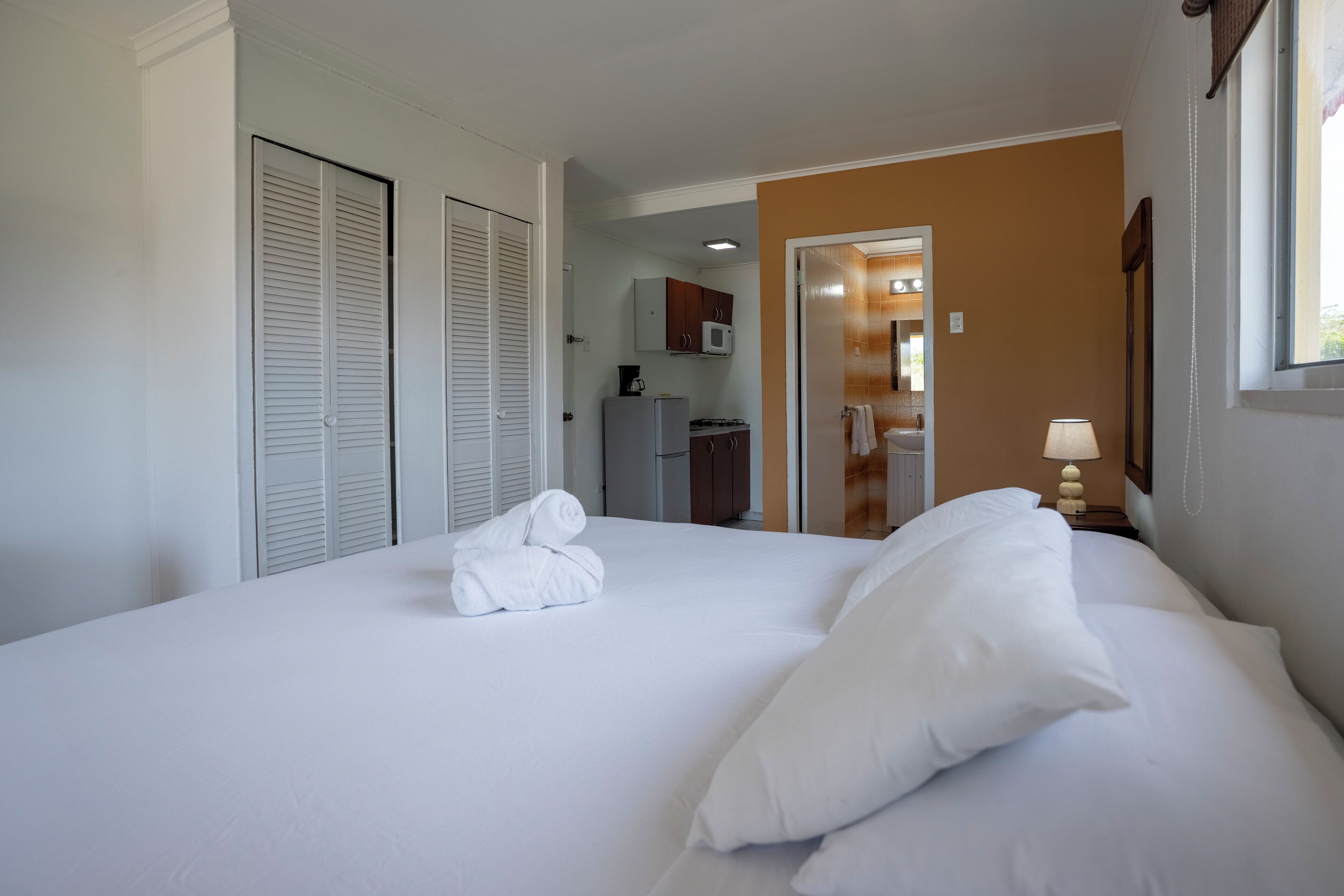 A hotel room with a neatly made bed, a bathroom with a mirror, and a kitchenette area.