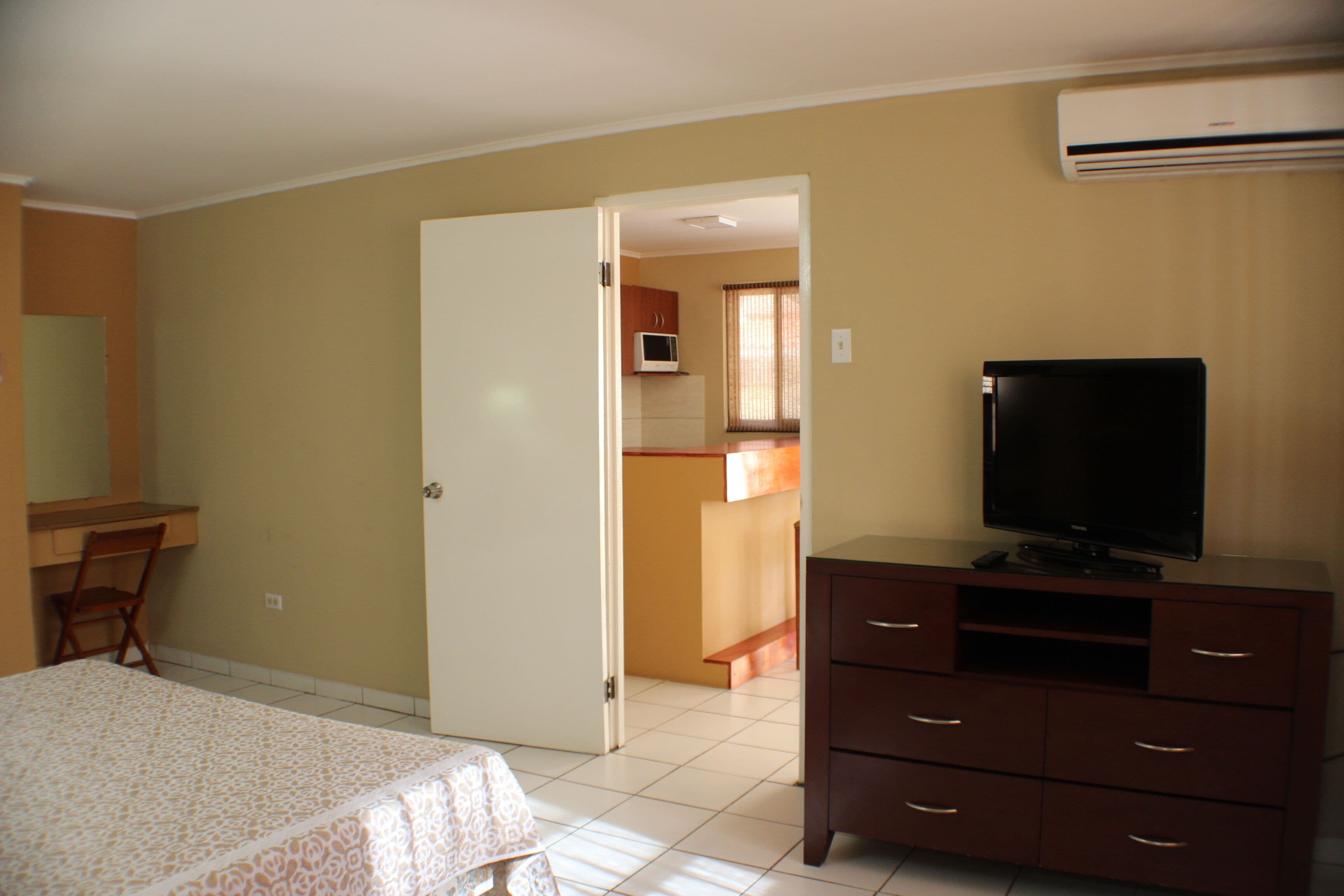 Un dormitorio con cama, cómoda, televisor y una puerta abierta que da a una zona de cocina.