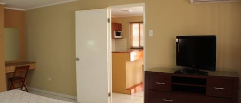 Un dormitorio con cama, cómoda, televisor y una puerta abierta que da a una zona de cocina.