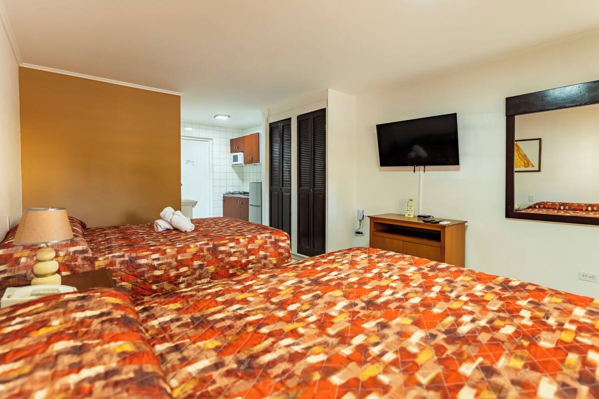 A hotel room with two beds, a TV, a desk, and a view of the kitchen.