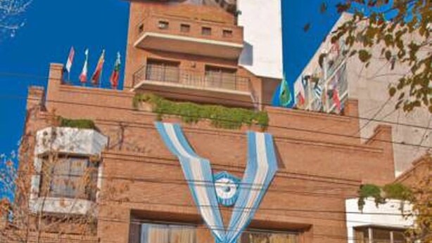 A hotel with a prominent entrance, a large banner with a logo, and multiple flags on the roof. - Reina Victoria Suites & Towers (Mendoza)