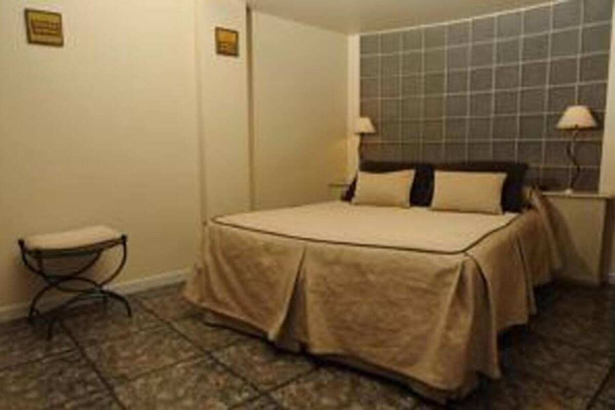 A hotel room with a bed, a nightstand, a lamp, and a small stool.