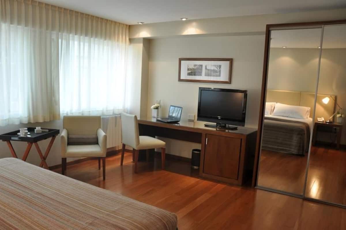 A hotel room with a bed, a desk with a computer, a chair, a TV, and a mirror.