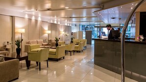 Un hall d’hôtel moderne comprenant un comptoir d’accueil, un espace salon avec des canapés et des fauteuils, et une grande fenêtre offrant une vue sur l’extérieur.