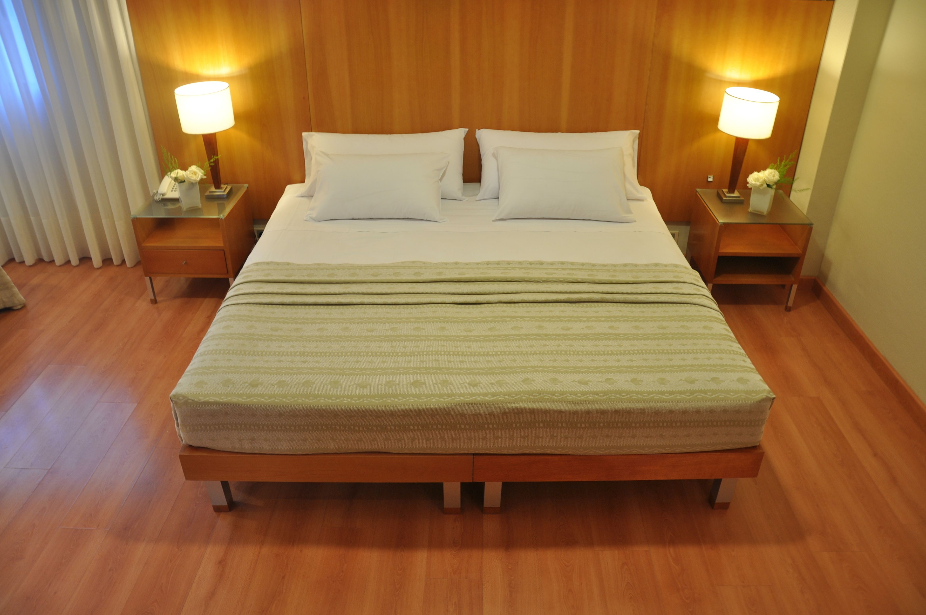 Cama bem arrumada com duas almofadas e um cobertor, com duas mesas de cabeceira laterais com luminárias e arranjos de flores.