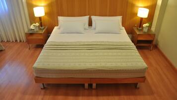Cama bem arrumada com duas almofadas e um cobertor, com duas mesas de cabeceira laterais com luminárias e arranjos de flores.