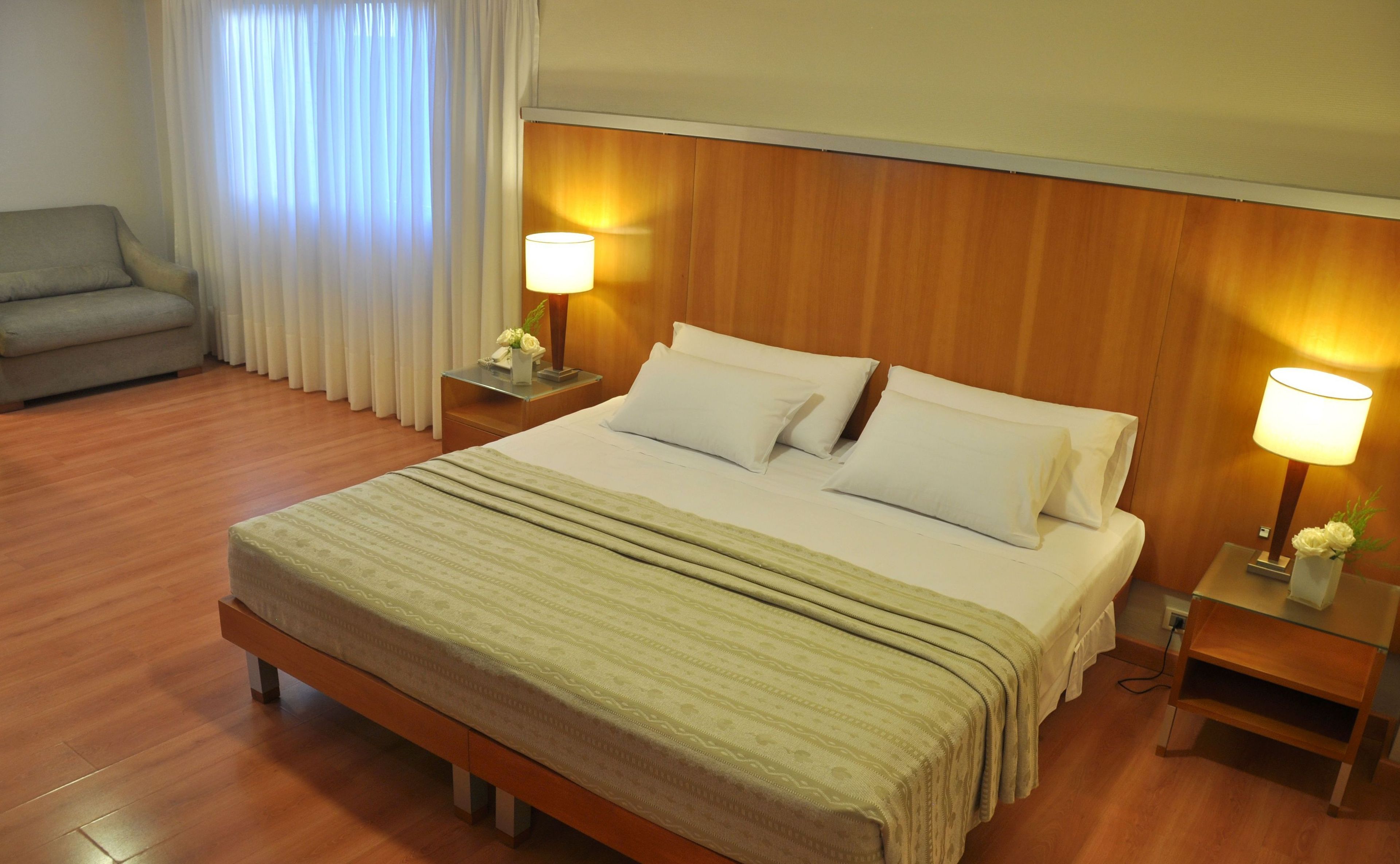 Una habitación de hotel con una cama grande, dos lámparas de noche, una mesita de noche, una silla y un ventanal con cortinas.