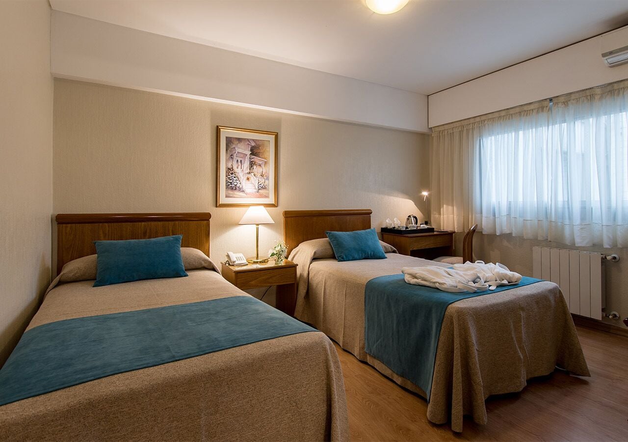 A hotel room with two beds, a nightstand with a lamp, a framed picture on the wall, and a window with curtains.