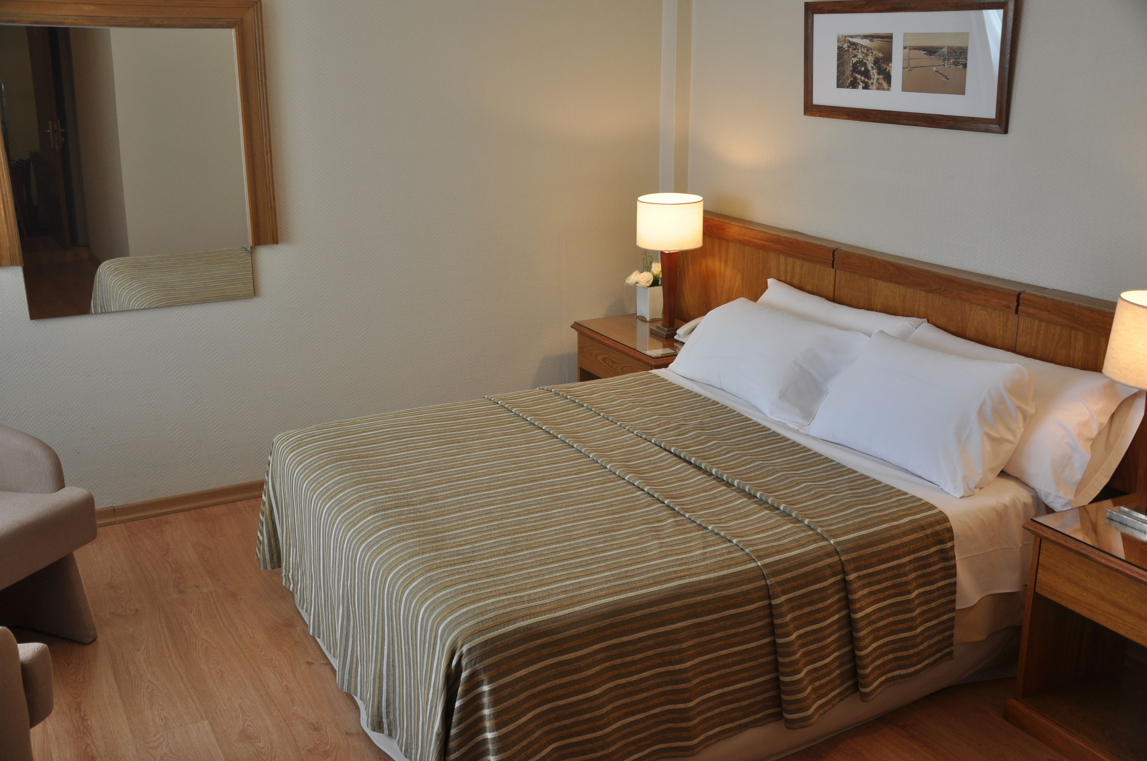 A neatly made bed with a striped bedspread, two bedside lamps, a wooden nightstand, a chair, and a framed picture on the wall.