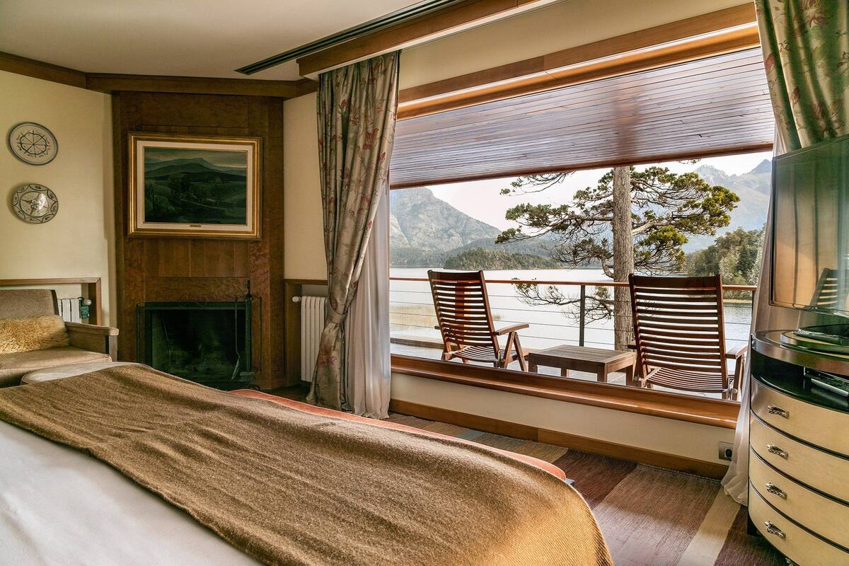 A bedroom with a large bed, a fireplace, a view of mountains and a lake through the window, and a wooden chair by the window.