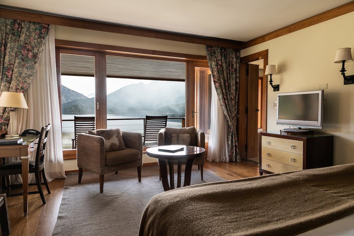 A hotel room with a large window offering a view of a lake and mountains, a television, a desk with a lamp, and two chairs.