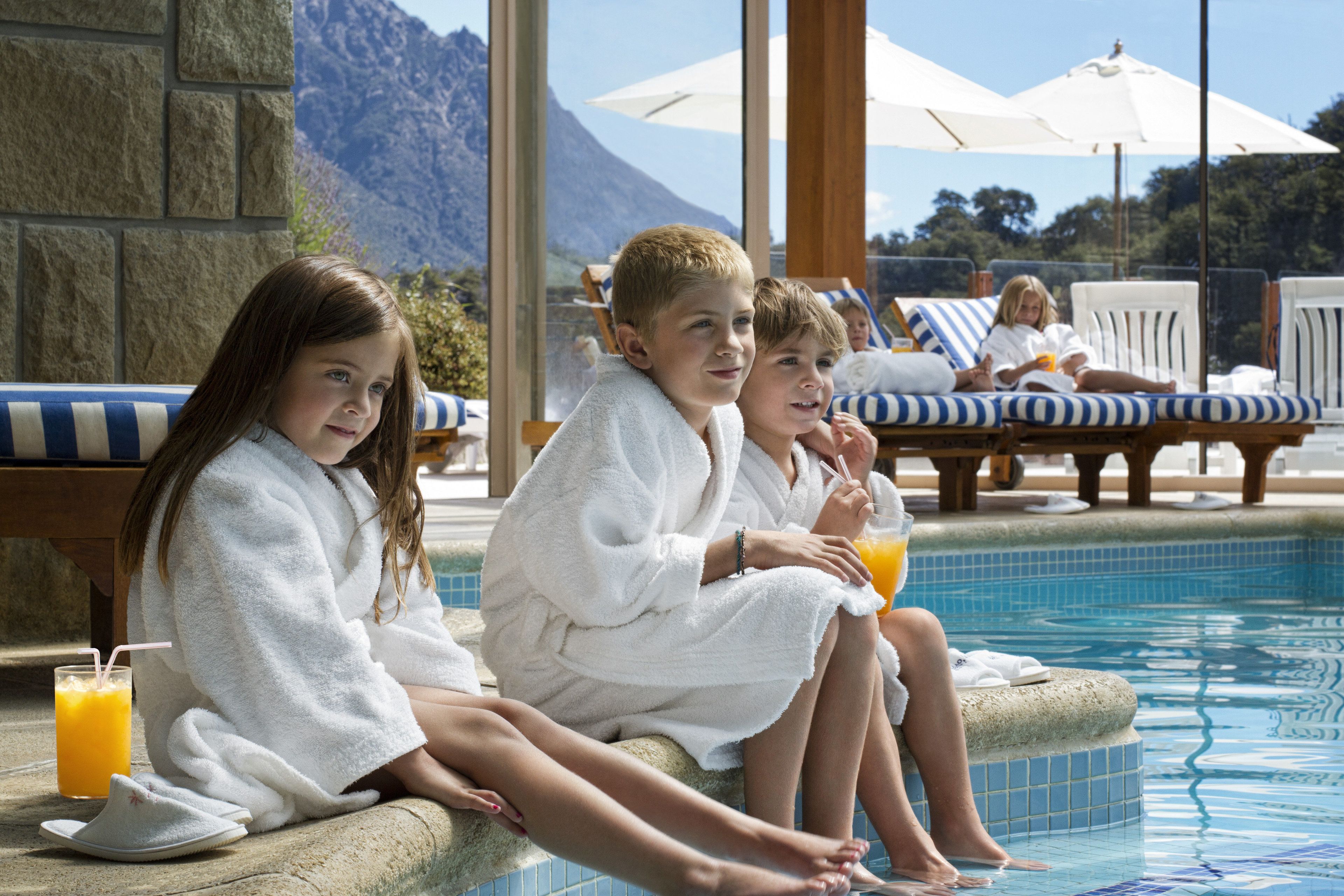 Three children in white bathrobes sitting by a pool with a mountain view in the background.