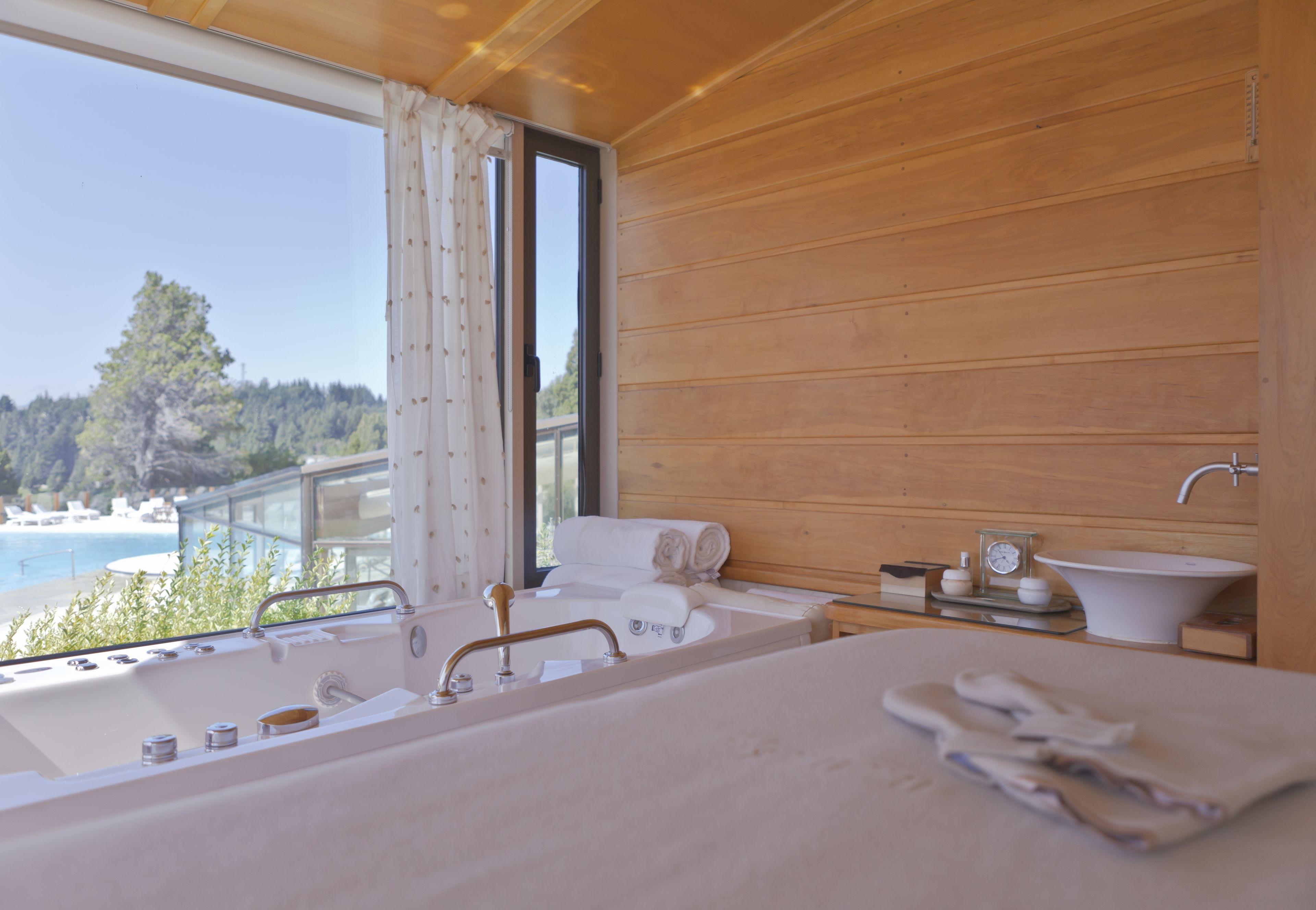 Ein Schlafzimmer einer Holzhütte mit großem Fenster, einer Badewanne, einem Waschbecken und Blick auf Strand und Wald.