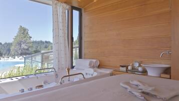 Ein Schlafzimmer einer Holzhütte mit großem Fenster, einer Badewanne, einem Waschbecken und Blick auf Strand und Wald.