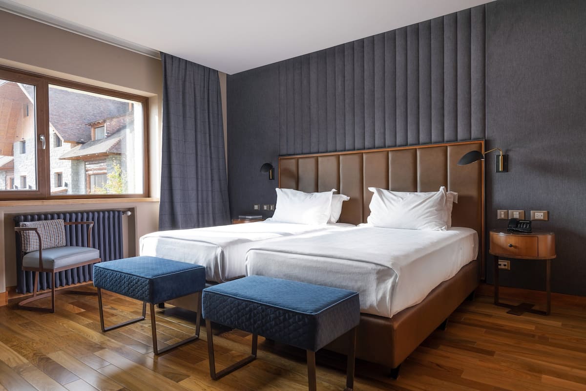A double room with two beds, a window, a radiator, a bedside table, and a small table with a chair.